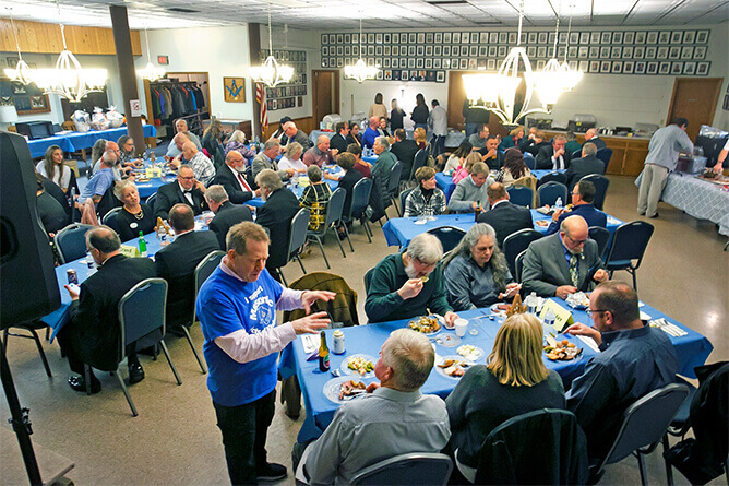 A large group of people sit at round tables covered with blue tablecloths, enjoying a meal in a well-lit banquet hall. Several individuals serve food from a buffet table at the back of the room.
