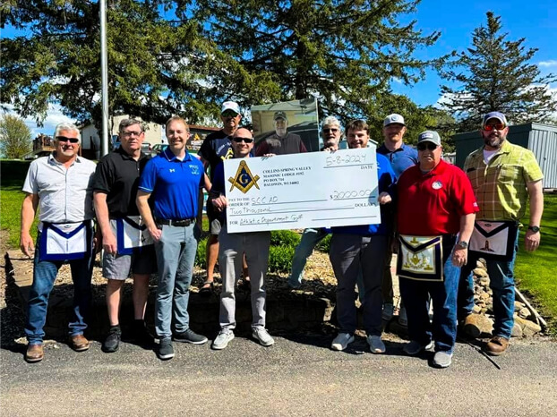 A group of ten men stand outdoors, several wearing Masonic aprons, holding a large ceremonial check for $2,000 made out to SCC HD. Trees and blue sky are visible in the background.