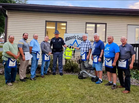 A group of men, including a police officer, stand in front of a building with a Masonic Lodge sign. Most are wearing Masonic aprons and one holds a defibrillator. They are outdoors on a grassy area.