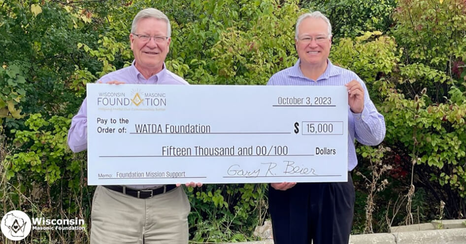 Two smiling men stand outdoors holding an oversized check for $15,000 made out to WATDA Foundation from the Wisconsin Masonic Foundation, dated October 3, 2023. Trees and greenery are in the background.