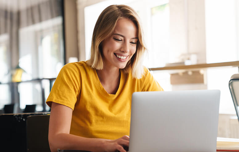 A young woman with blonde hair and a yellow t-shirt is smiling while working on a laptop in a bright, modern indoor setting.