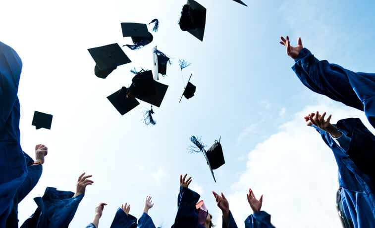 Graduates in blue gowns toss their caps into the air, celebrating outdoors under a bright sky. The photo is taken from a low angle, focusing on the flying caps and outstretched hands.
